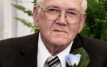 Elderly man in a suit and tie with a blue boutonnière, sitting in front of green plants, looking directly at the camera.
