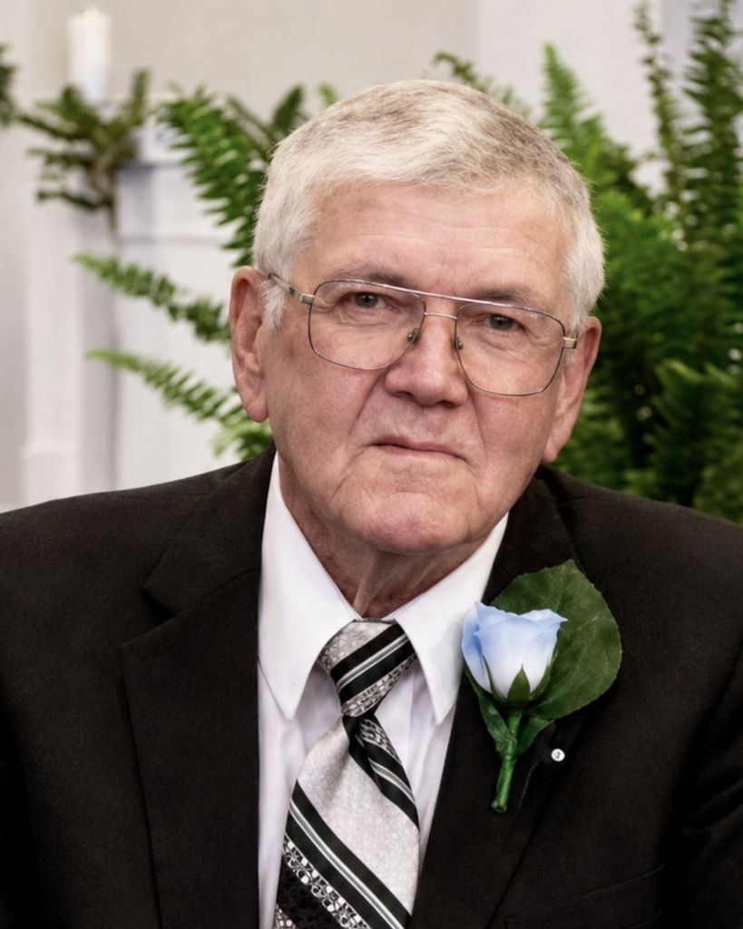 Elderly man in a suit and tie with a blue boutonnière, sitting in front of green plants, looking directly at the camera.