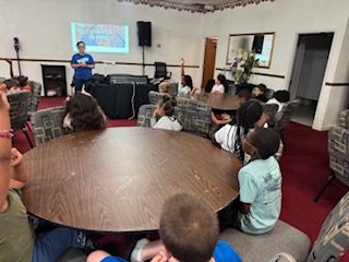 Children seated around tables in a classroom, listening to an instructor at the front with a projection screen behind them.