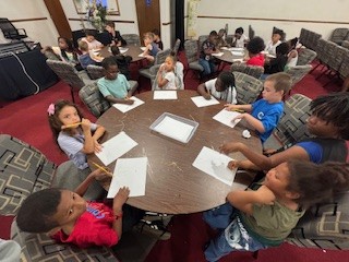Children sitting around tables in a classroom setting, drawing on white paper with pencils, appearing engaged and focused on their activities.