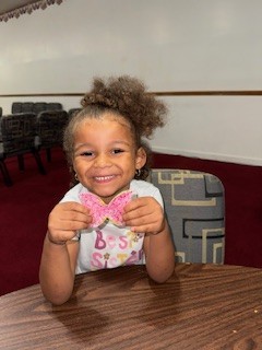 Smiling young girl sitting at a table, holding a pink frosted heart-shaped cookie. Empty chairs and maroon carpet are in the background.