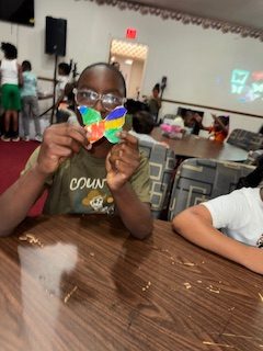 A child holding up a colorful paper butterfly while sitting at a table in a classroom with other children and decorations in the background.