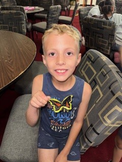 A young boy with blond hair smiles and shows a yellow and black butterfly patch while sitting on a patterned chair in a room with tables and chairs.