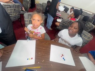 Two young girls sit at a table with blank papers and colored objects, participating in a group activity with other children in a classroom setting.