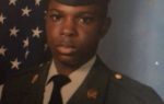 A young man in a military uniform poses in front of an American flag backdrop.