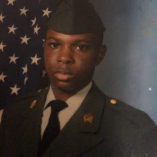 A young man in a military uniform poses in front of an American flag backdrop.