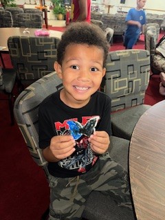 Smiling young boy sitting at a table, holding a colorful craft butterfly, inside a room with patterned chairs and other children in the background.