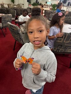 A young girl in a gray hoodie holds an orange craft butterfly, smiling at the camera inside a room where other children are seated and working.