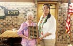 Two women standing indoors, one holding a plaque with engraved nameplates. There is an American flag and a brick wall with a "Loft" sign in the background.