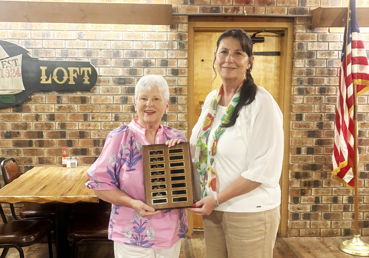 Two women standing indoors, one holding a plaque with engraved nameplates. There is an American flag and a brick wall with a "Loft" sign in the background.