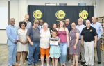 A group of people poses indoors, with a young girl holding a certificate at the center, in front of a black and yellow banner.