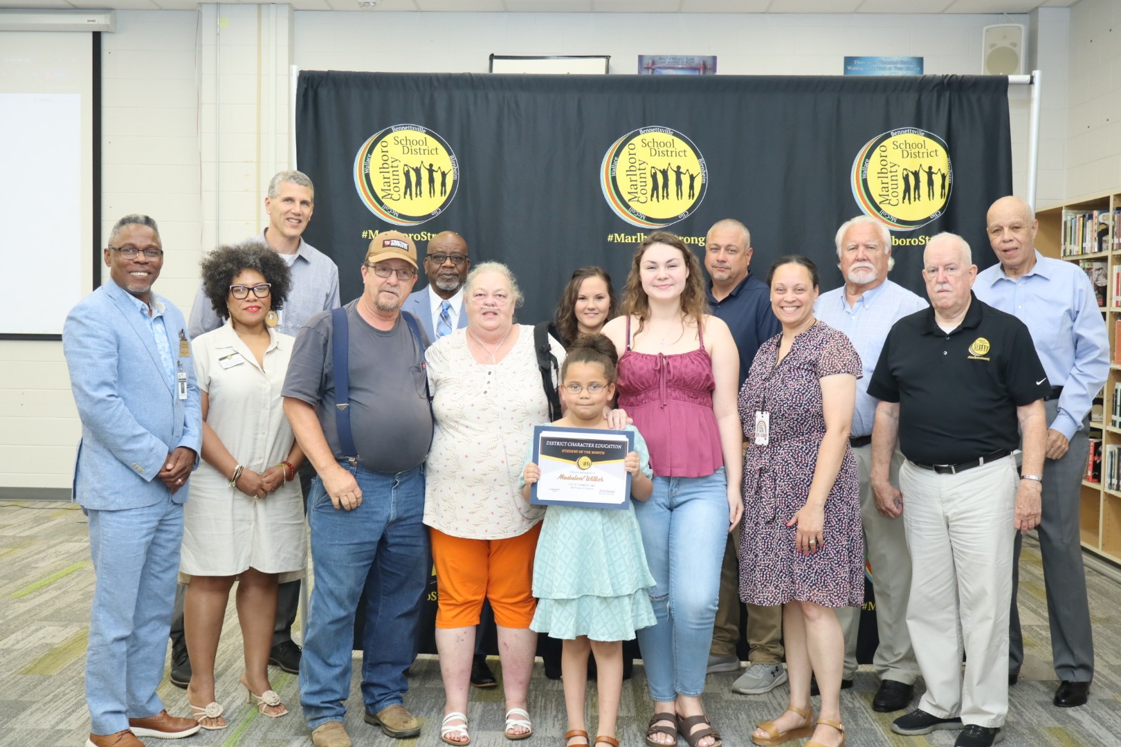 A group of people poses indoors, with a young girl holding a certificate at the center, in front of a black and yellow banner.
