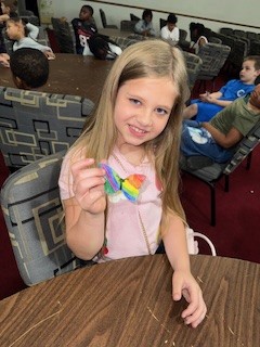 Young girl sitting at a table in a classroom holds up a colorful butterfly craft and smiles at the camera.