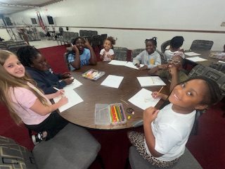 A group of children sit around a table drawing with colored pencils and smiling in a classroom or community center setting.
