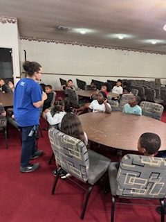 A group of children sitting at round tables in a room with red carpet, listening to an adult standing and speaking to them.