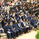 Graduates in blue caps and gowns sit in rows during a graduation ceremony, with an audience watching in the background.