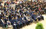 Graduates in blue caps and gowns sit in rows during a graduation ceremony, with an audience watching in the background.