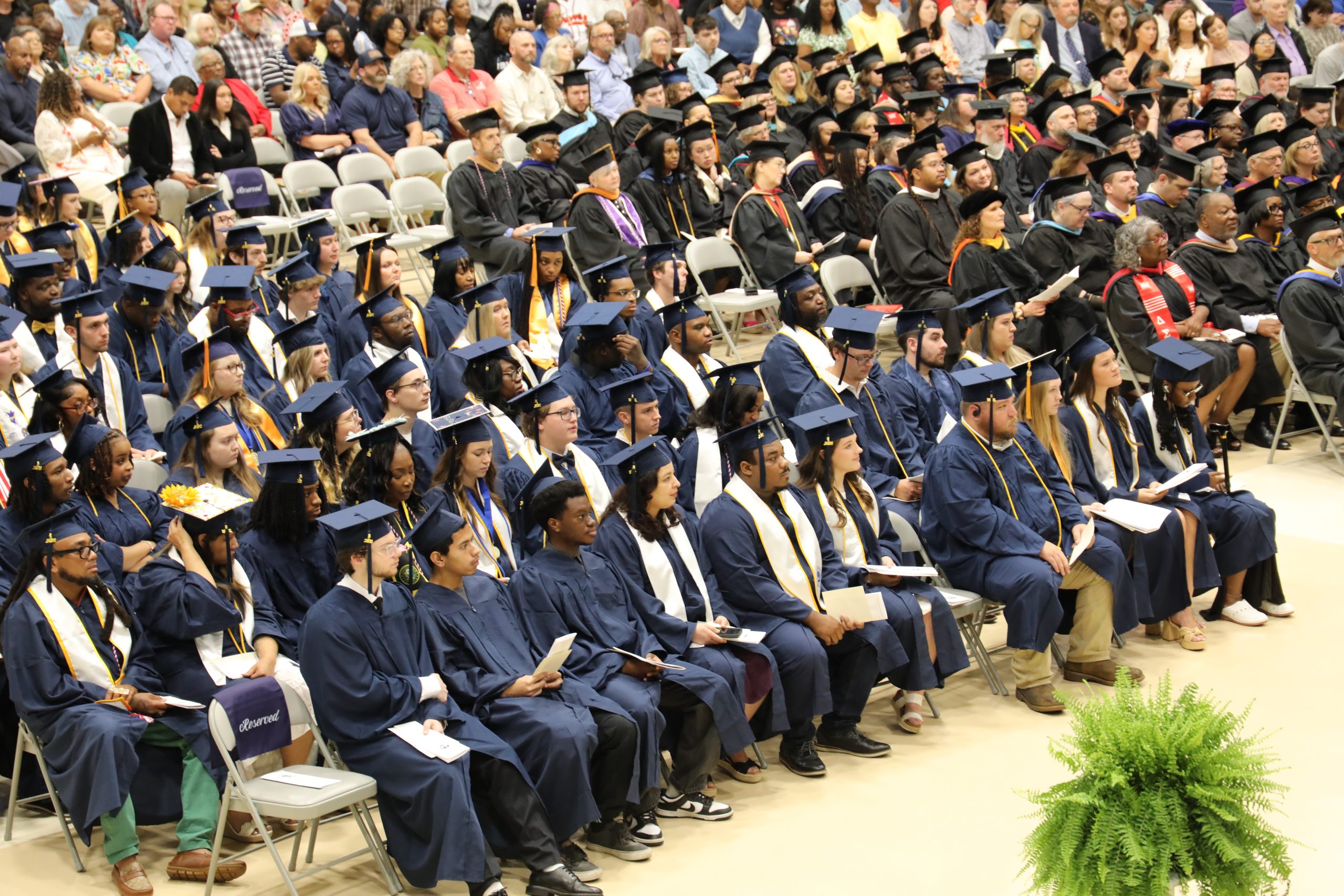 Graduates in blue caps and gowns sit in rows during a graduation ceremony, with an audience watching in the background.