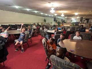 Children sitting at round tables in a large room with red carpet, some raising hands and looking forward, possibly attending a class or event.