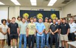 A group of men pose for a group photo indoors in front of a black and yellow banner.
