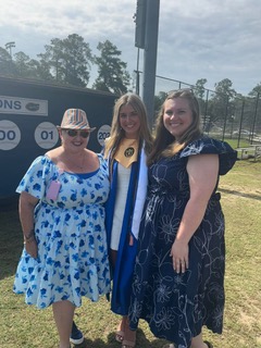 Three women smiling outdoors, one in a graduation gown and cap, standing between two women in floral dresses on a sunny day near a sports field.