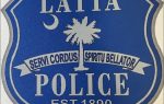 Blue Latta Police badge with a crescent, palmetto tree, Latin motto "Servi Cordus Spiritu Bellator," and "EST. 1890" at the bottom.