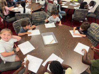 Children sitting around tables drawing with pencils on white paper in a classroom setting.