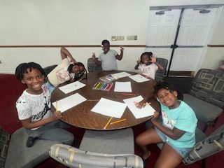Five smiling children sit around a table with drawing supplies, posing and making peace signs while starting artwork on white paper in a brightly lit room.