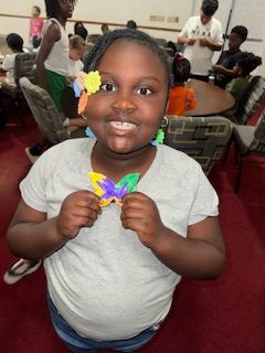Smiling girl holds a colorful butterfly craft, standing in a room with other children sitting and working at tables in the background.