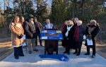 A group of people gathered outdoors for an unveiling ceremony, standing around a historical marker display, with trees and winter clothing indicating a cold day.