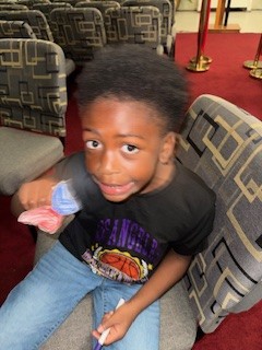 Young boy sitting on a patterned chair, holding candy, wearing a black Los Angeles t-shirt and jeans, with seats and red carpet in the background.