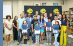Group of adults and children posing indoors, some children holding certificates and artwork in front of a black and yellow banner, suggesting an award or recognition event.