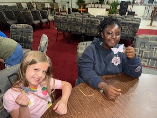 Two girls sit at a table in a room with chairs, smiling and holding up craft butterflies they made.