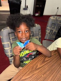 Smiling child sitting at a table, holding up a handmade blue and white butterfly craft, in a room with patterned chairs and a red carpet.