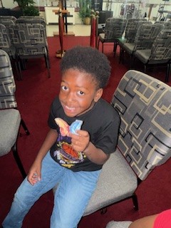 Smiling boy sitting on a chair in a room with patterned seats, holding colorful snacks in his hand.