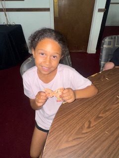 Smiling young girl with curly hair holding a butterfly-shaped cookie, sitting at a brown table in a room with maroon carpet and simple decor.