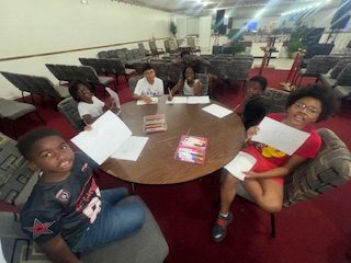 Seven children sit around a table in a room, holding up papers and smiling, with colored pencils and markers in the center.