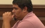 Man in a pink polo shirt sitting in a courtroom, resting his face on his hand, appearing thoughtful or concerned.