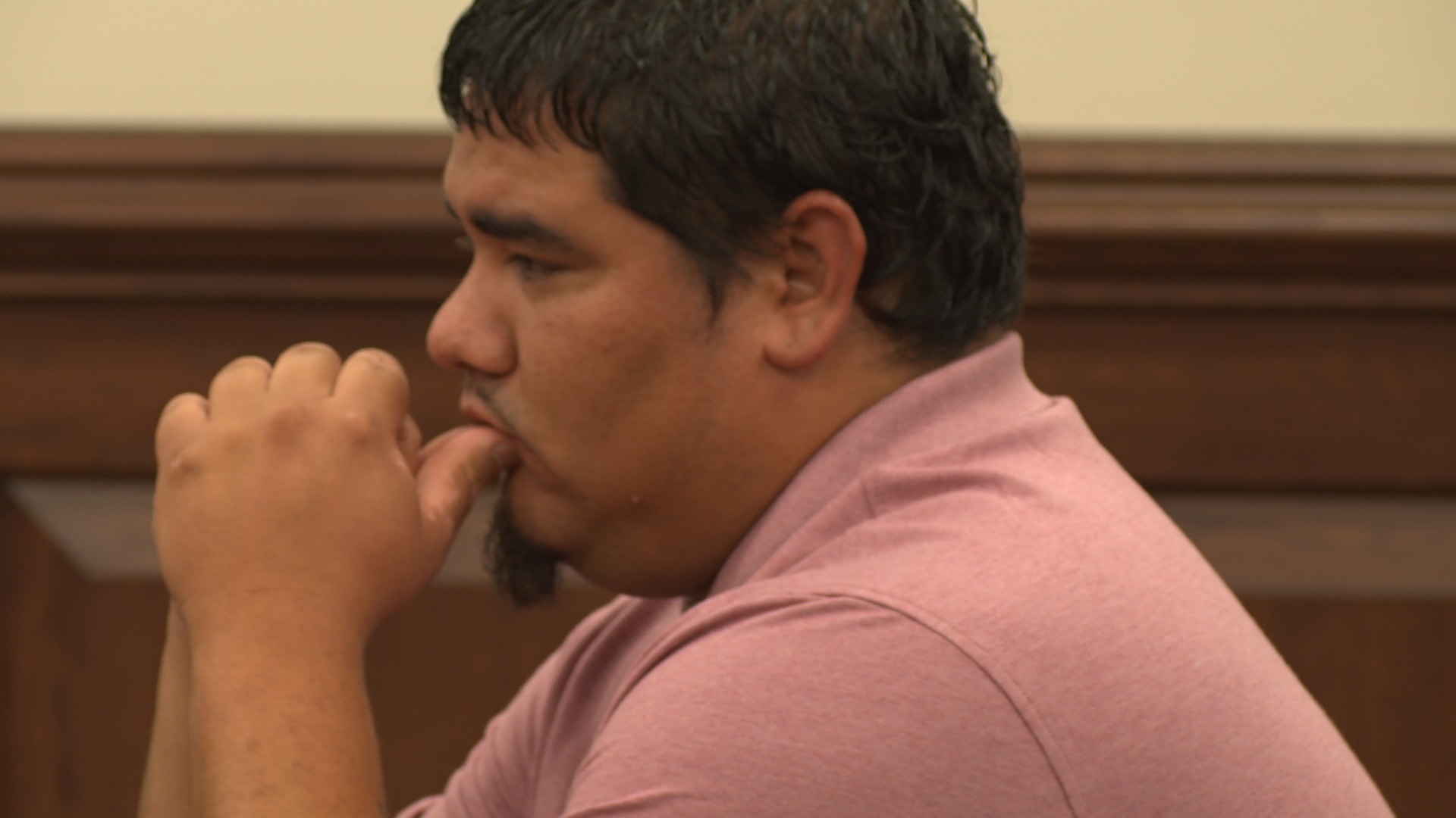 Man in a pink polo shirt sitting in a courtroom, resting his face on his hand, appearing thoughtful or concerned.