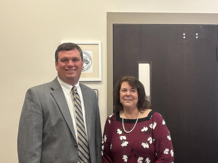 A man in a gray suit and a woman in a patterned blouse standing indoors, smiling at the camera.