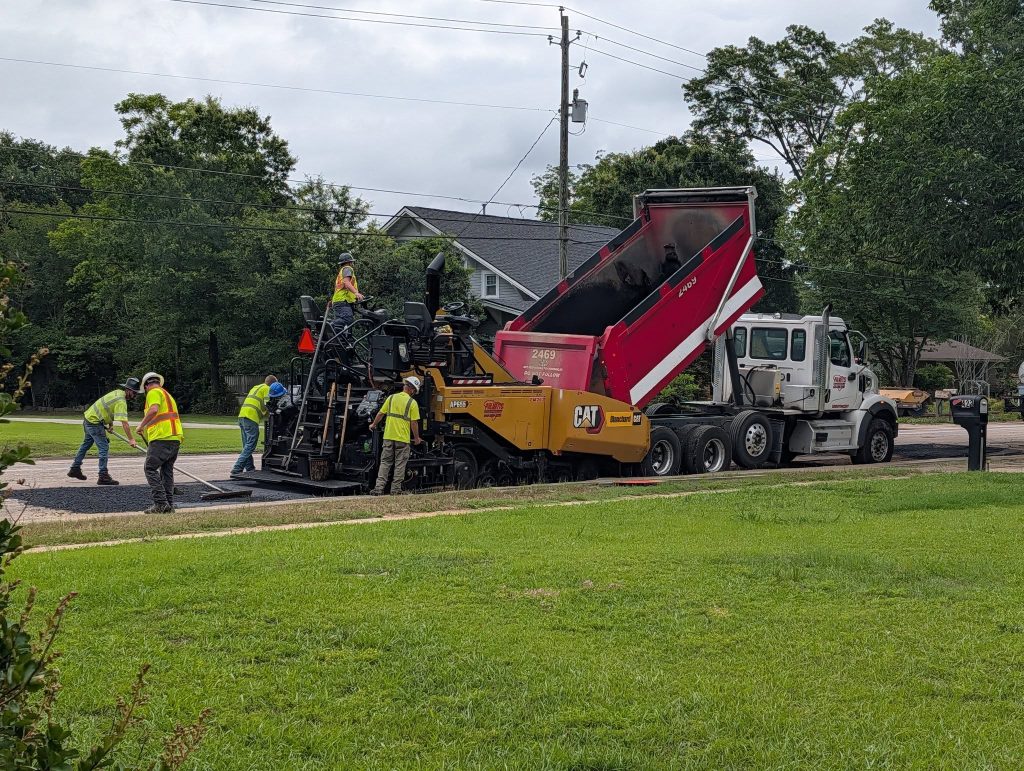 Workers paving a road using construction machinery and a dump truck on a cloudy day, with trees and a house in the background.