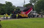 Workers paving a road using construction machinery and a dump truck on a cloudy day, with trees and a house in the background.