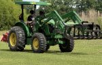 Two people ride a green tractor with a front loader attachment on a grassy field, surrounded by trees and bushes under a clear sky.