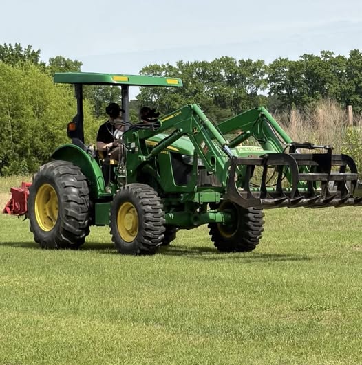 Two people ride a green tractor with a front loader attachment on a grassy field, surrounded by trees and bushes under a clear sky.