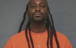 A man with long dreadlocks wearing an orange shirt stands against a gray background, looking at the camera with a slight smile.