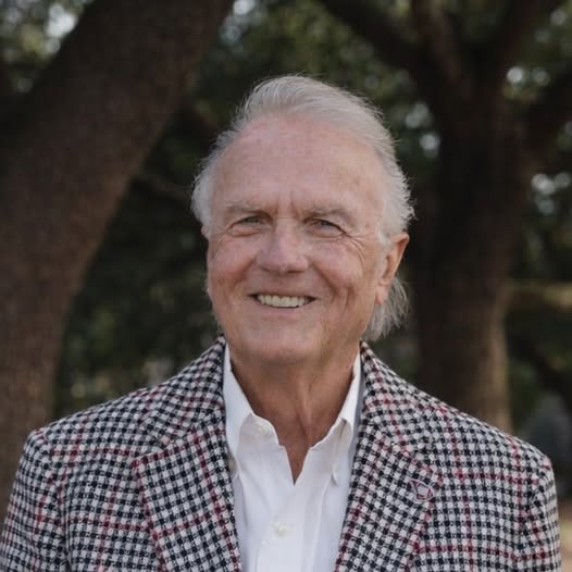 older man with gray hair, wearing a checked blazer and white shirt, smiles outdoors with trees in the background.