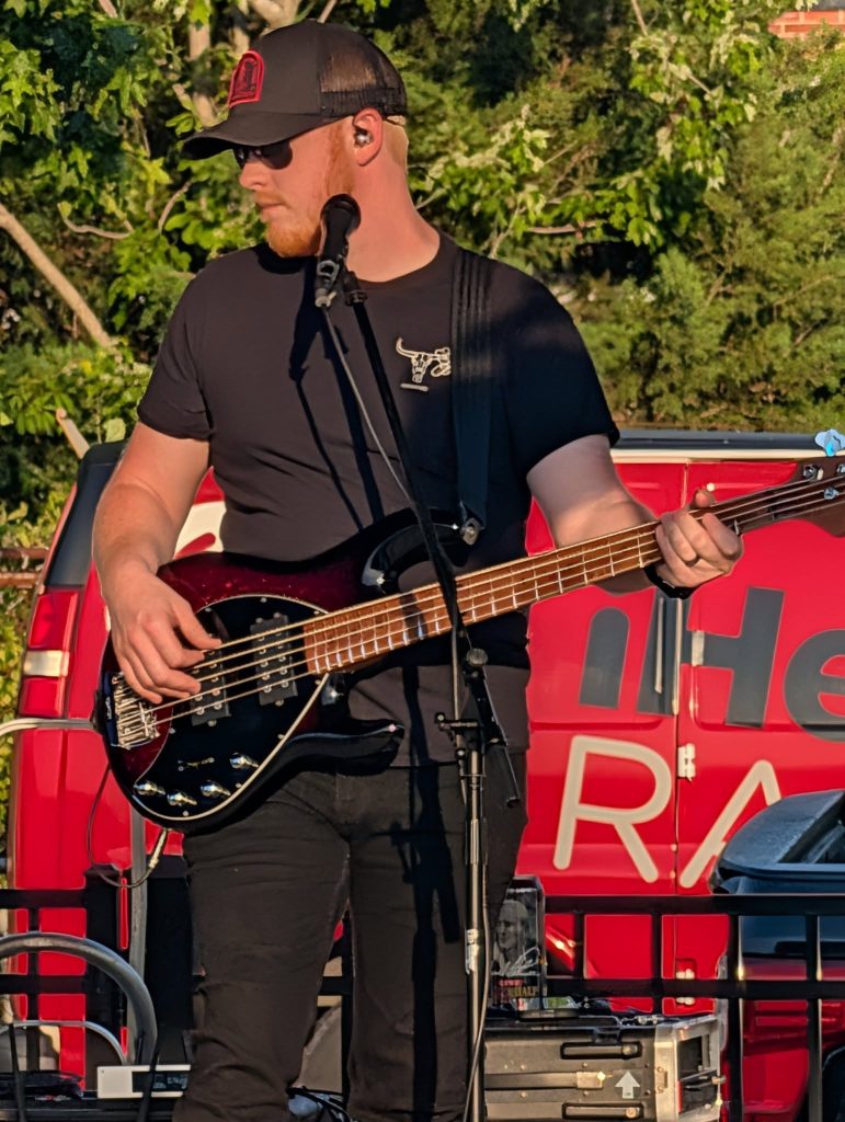 Man in black shirt and hat playing electric bass guitar outdoors, with a red van and trees in the background.