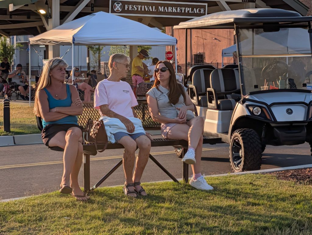 Three women sit on a bench outdoors, talking, with a golf cart, tents, and a "Festival Marketplace" sign visible in the background.