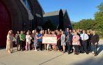 A group of people stands outside a brick building, holding a large ceremonial check, with clear blue sky and trees in the background.
