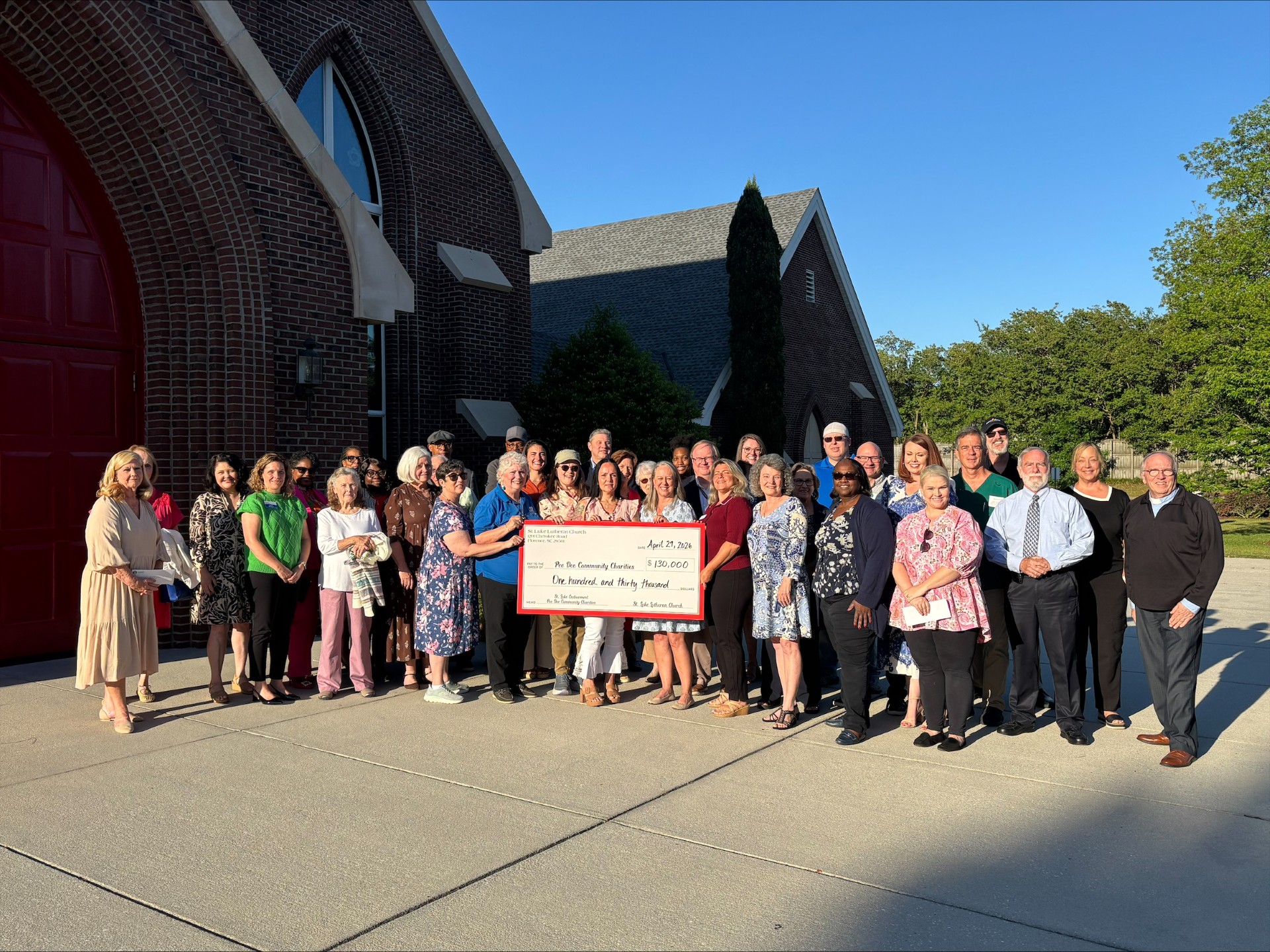 A group of people stands outside a brick building, holding a large ceremonial check, with clear blue sky and trees in the background.